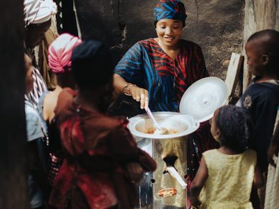 African woman cooking surrounded by cheerful children outdoors in a village setting.